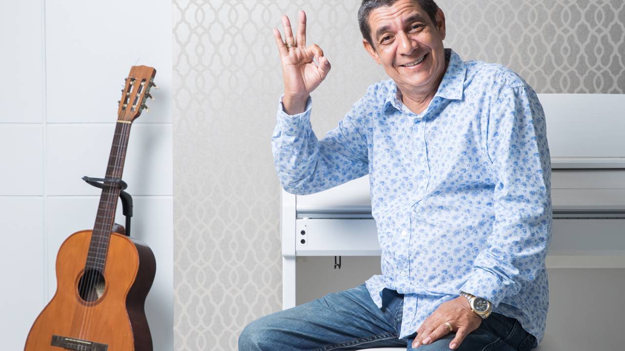 Zeca Pagodinho sentado num banco branco, com um violão ao lado dele, fazendo sinal de ok com a mão