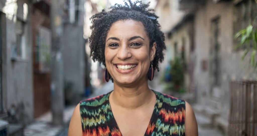 marielle franco marielle franco está sorrindo. Ela usa vestido preto com stampas verdes e vermelhas. O cabelo está semipreso.