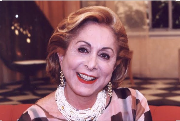 Aracy Balabanian é uma mulher branca de cabelos claros. Está usando batom vermelho e um volumoso colar branco.