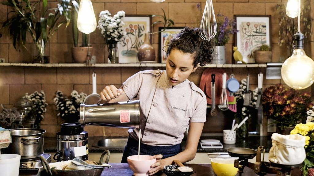 Conflor – Leite Vegetal – Confeiteira Luísa Mendona – Foto Klacius Ank 2.jpg A imagem mostra a confeiteira vegana Luisa Mendonça na cozinha, despejando numa vasilha o leite vegetal que está no copo de um liquidificador