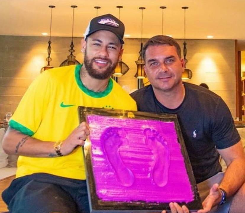 A imagem mostra o jogador de futebol Neymar sentado ao lado de Flávio Bolsonaro; ambos seguram o molde com a marca de seus pés, que entraram na calçada da fama do Maracanã