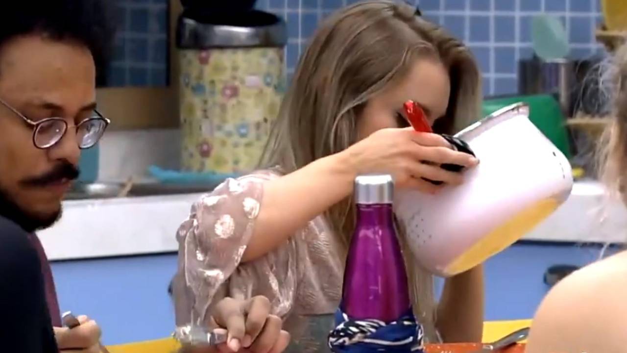 A imagem mostra a atriz Carla Diaz sentada à mesa, segurando uma panela e colocando o rosto bem próximo a ela