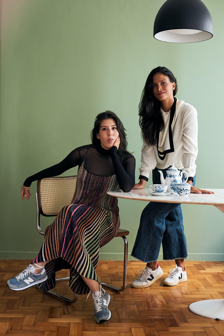 Duas mulheres brancas com cabelos longos castanhos posam na foto, uma em p&eacute; com blusa branca, outra sentada com blusa preta