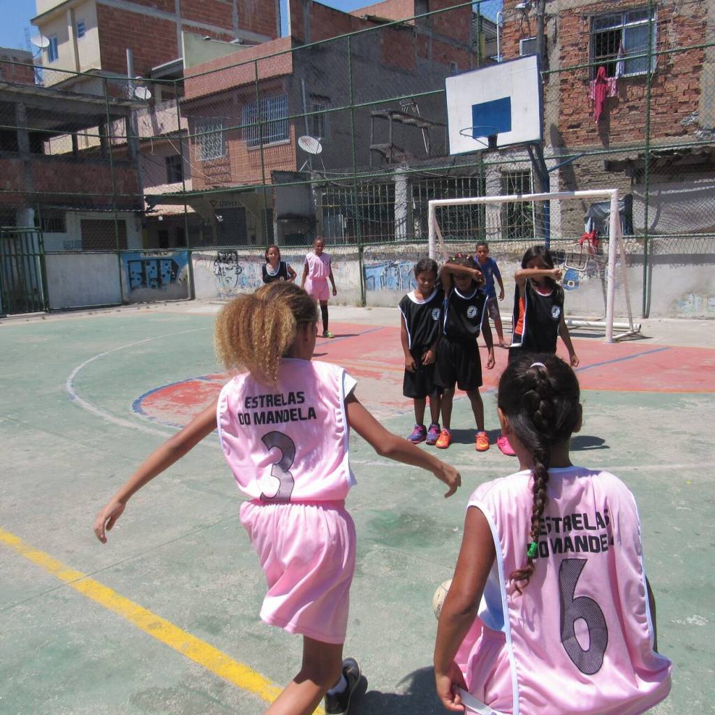 Foto 2 – Divulgação Imagem mostra meninas com uniforme rosa claro jogando futebol em uma quadra da comunidade