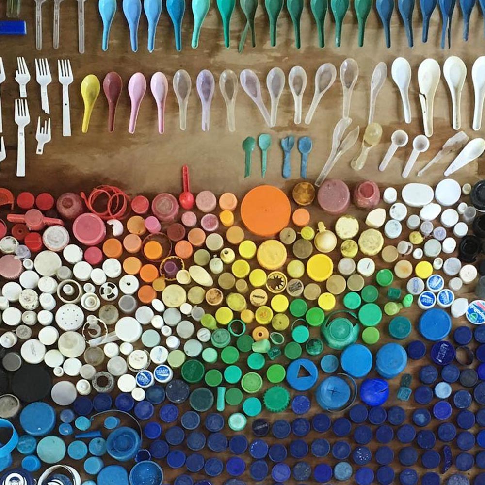 Foto mostra diversos tipos de pl&aacute;stico, como colheres descart&aacute;veis e tampas pl&aacute;sticas de diversas cores em uma mesa, uma ao lado da outra