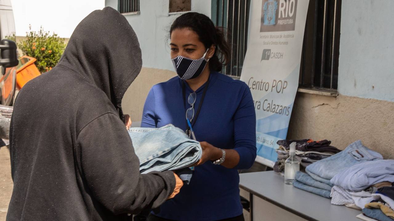 Foto mostra pessoa entregando roupa a pessoa em situação vulnerável