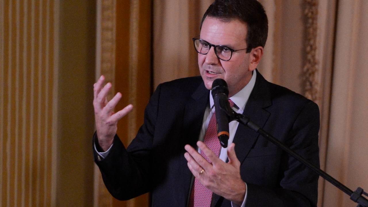 O prefeito do Rio de Janeiro, Eduardo Paes fala durante cerimônia de posse do secretariado. Ele está gesticulando com as mãos e usa óculos. Está vestindo paletó escuro com camisa clara e gravata avermelhada.