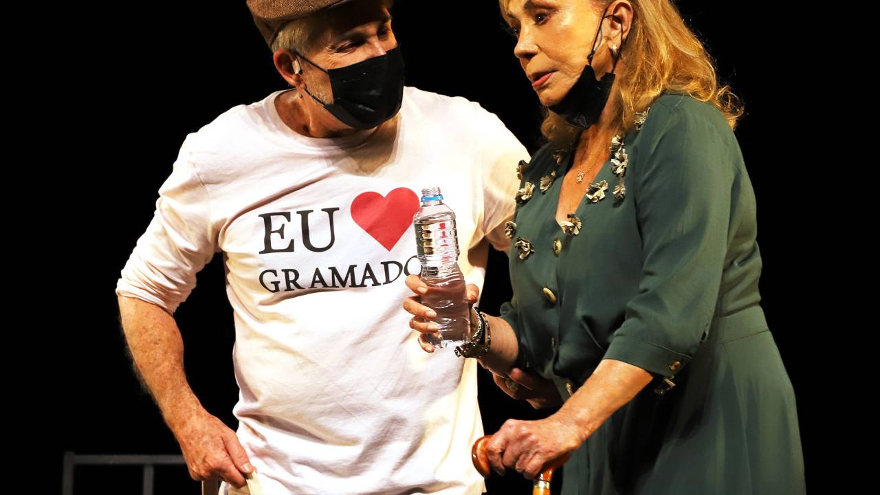 Arlete Salles com um vestido verde segura uma bengala e uma garrafa de água ao lado de Edwin Luisi, de máscara preta e uma blusa com os dizeres 'Eu Amo Gramado'