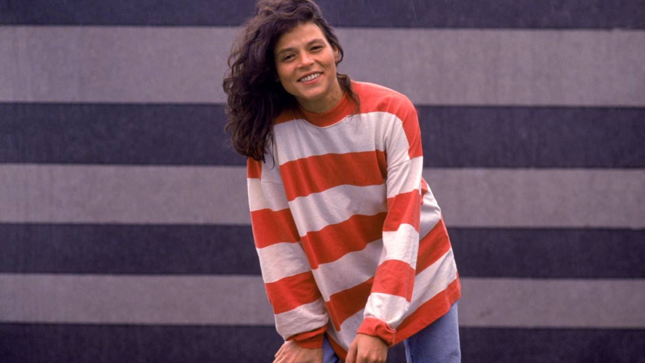 A cantora Cássia Eller está sorrindo. Ela está com os cabelos compridos jogados sobre seu ombro direito. Ela veste moletom vermelho e branco, listrado na horizontal, e calça jeans clara. A mão esquerda segura um cigarro e a mão direita está apoiada no joelho direito.