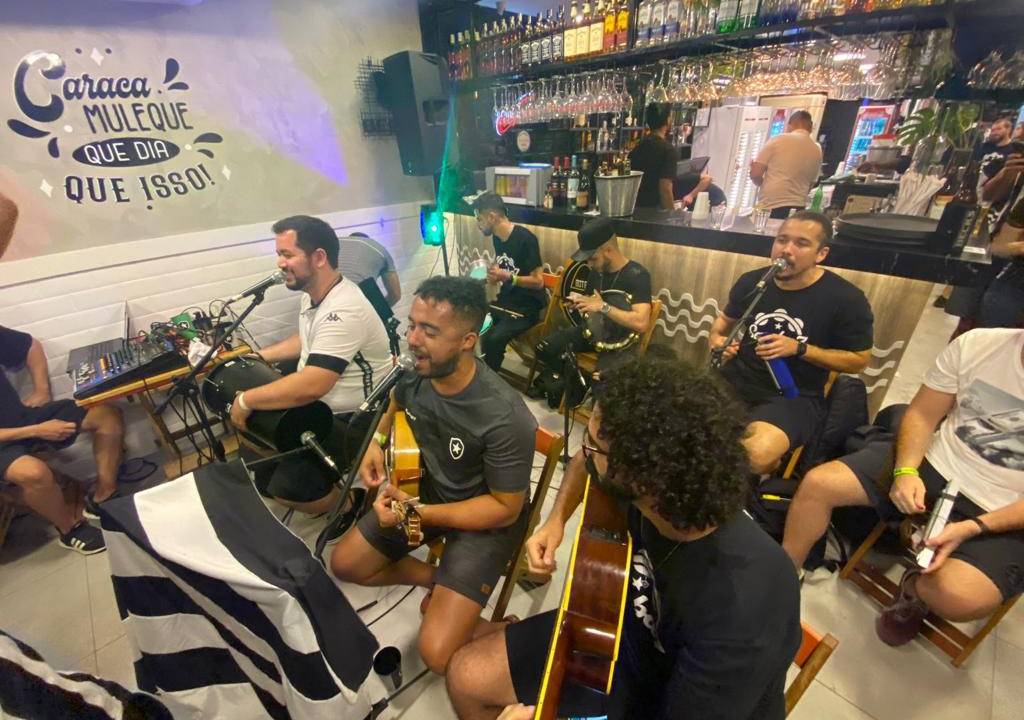 Grupo de música se apresenta dentro de um bar. Os sete integrantes, três na frente e quatro atrás, estão sentados e com microfones à frente. O piso do bar é preto e banco.