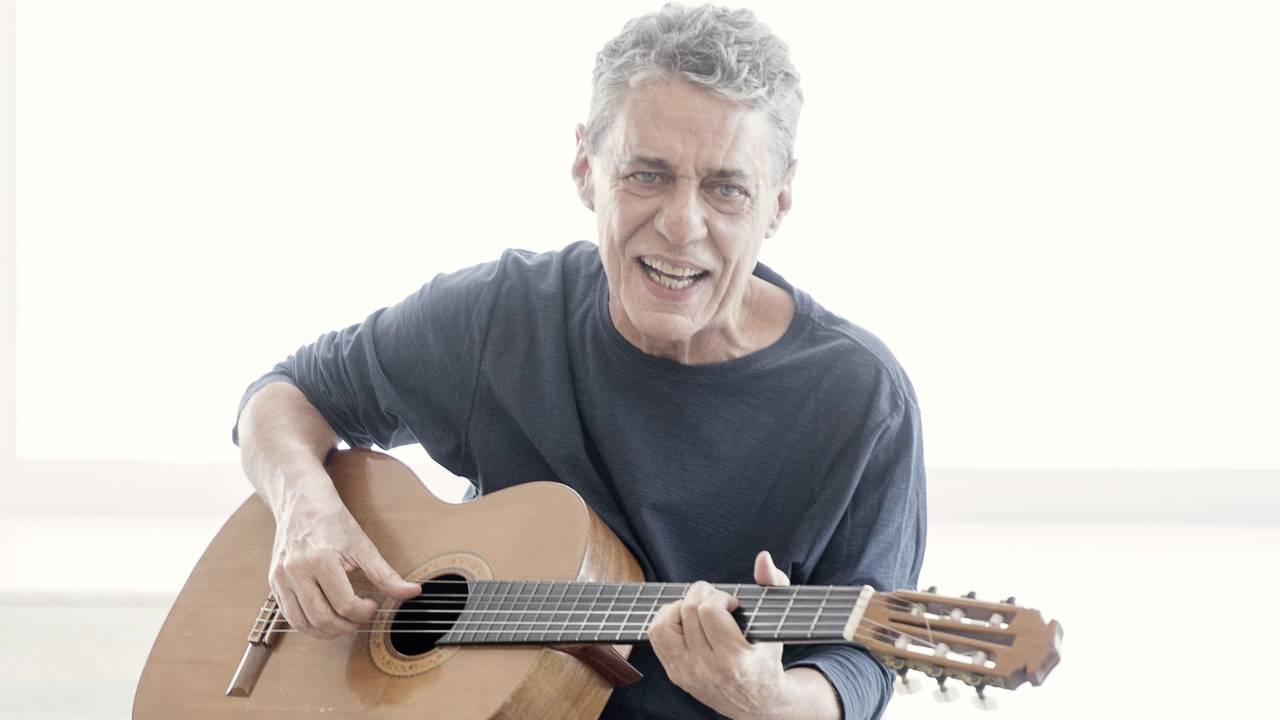 O cantor e compositor Chico Buarque está sentado em pose de quem toca o violão e com a boca entreaberta. Ele veste blusa azul-escuro e apoia o violão em um dos joelhos.
