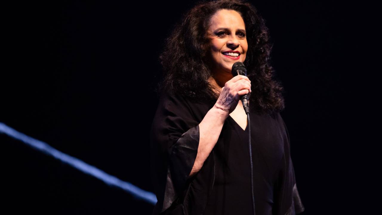 A cantora Gal Costa de vestido preto no palco com fundo preto. Ela segura o microfone e está sorrindo.