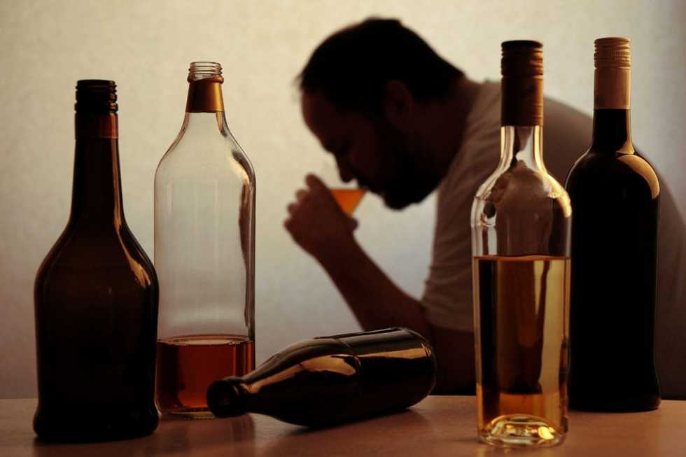 Homem sentado diante de garrafas de bebidas alcoólicas.
