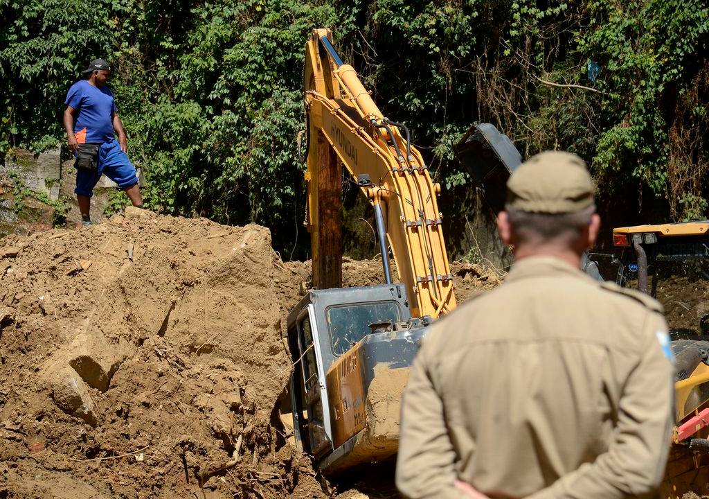 Foto mostra escavadeira tirando entulhos de desabamento com monitoramento de um bombeiro