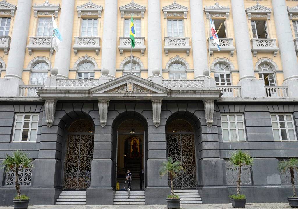 Foto da fachada do Museu da Justiça do Rio de Janeiro, retangular e simétrica, em estilo eclético classicizante, , com tendências ao neorrenascentismo italiano, do final do século XIX. O pórtico de entrada é formado por três grandes portões em ferro ricamente trabalhado em arabescos. Acima do pórtico e do segundo pavimento até a base do quinto, uma sequência de oito colunas confere grandeza à fachada principal.
