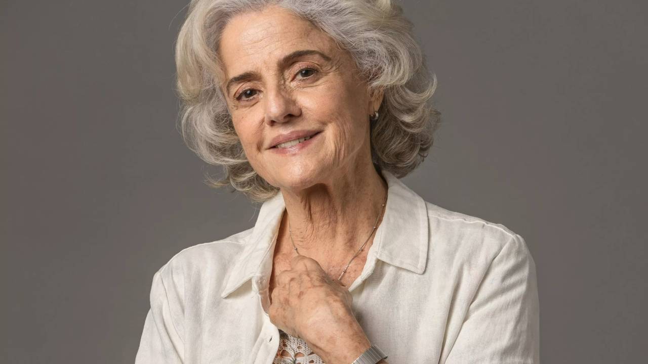 A atriz Marieta Severo de cabelos brancos. Ela usa camisa clara e traz a mão esquerda, no colo, além de usar relógio no pulso esquerdo.