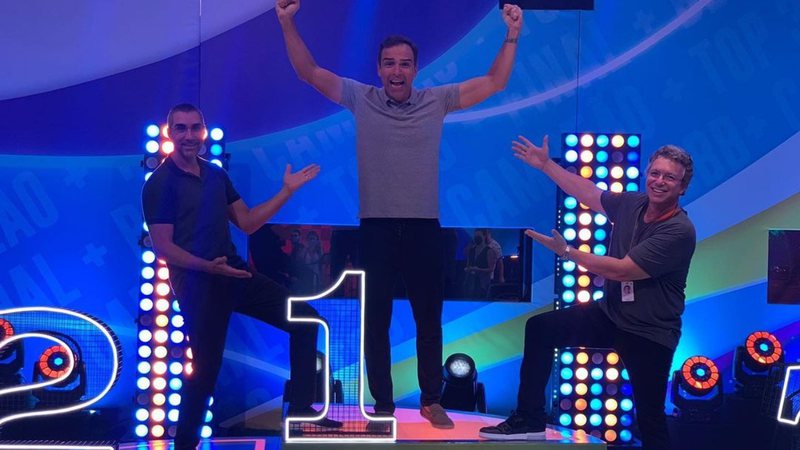 Tadeu Schmidt no alto do pódio do BBB, com o diretor Rodrigo Dourado no segundo lugar e o diretor Boninho no terceiro, os dois com os braços na direção de Tadeu.