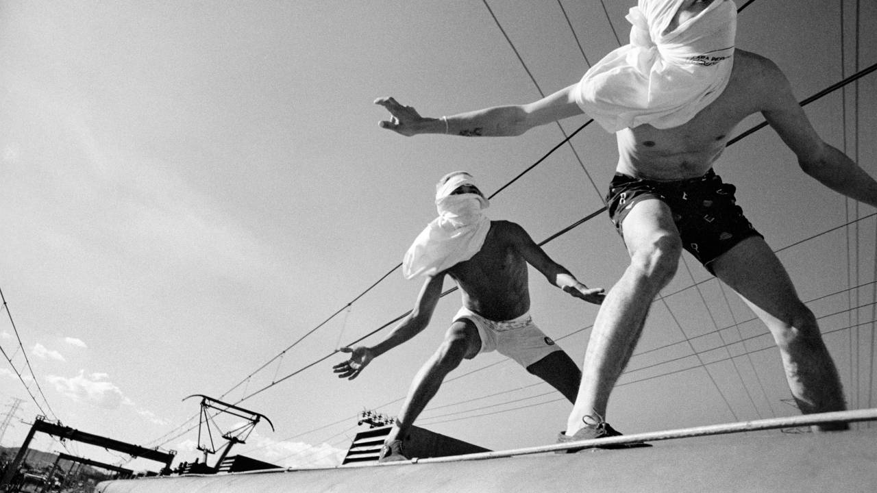 Foto em preto e branco de dois jovens com a cabeça cobertura com camisetas "surfando" um trem -