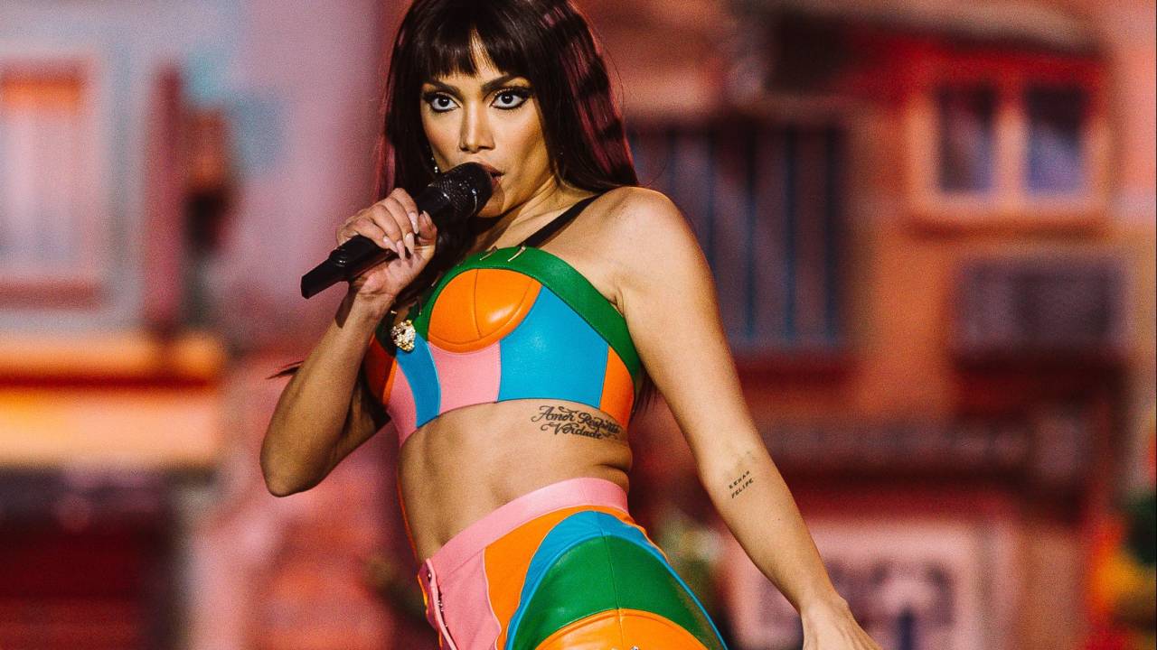 Anitta com top e hot pants nas cores verde, laranja, rosa e azul-turquesa. Ela está no palco cantando.