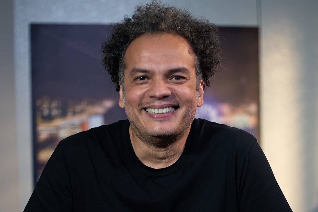 Zé Ricardo sentado, sorrindo, de blusa preta