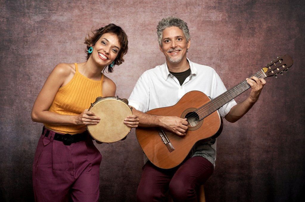 Gabriel Sant’Anna e Martha Paiva 1 – fotógrafo – João Saidler Foto mostra mulher segurando pandeiro e sorrindo ao lado de homem segurando violão e sorrindo