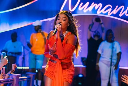 Foto mostra cantora Ludmilla usando vestido laranja e cantando com microfone