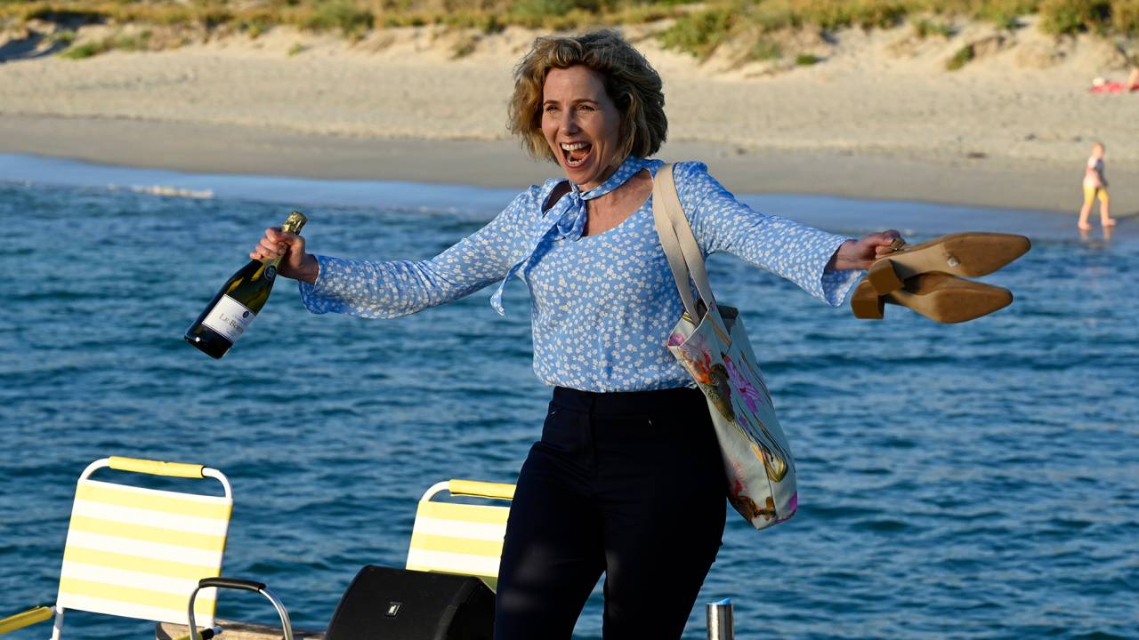 Sally Philips tem cabelos louros e médios. Ela estáde calça preta e camisa azul, está com os braços abertos, segurando uma garrafa de vinho com a mão direita e com a boca aberta em expressão de felicidade. Ela usa uma bolsa clara no ombro esquerdo. Ao fundo, o mar.