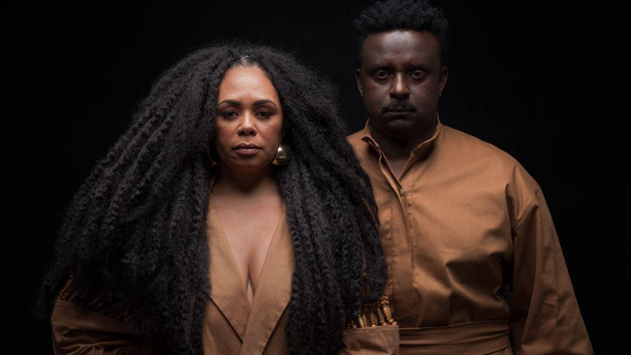 FOto mostramulher e homem negros usando roupa bege e na frente de um fundo preto