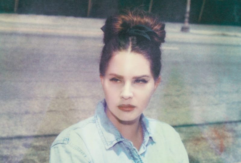 Lana del Rey está de camisa social azul e usa coque no alto da cabeça. Ela tem o cabelo liso e escuro.