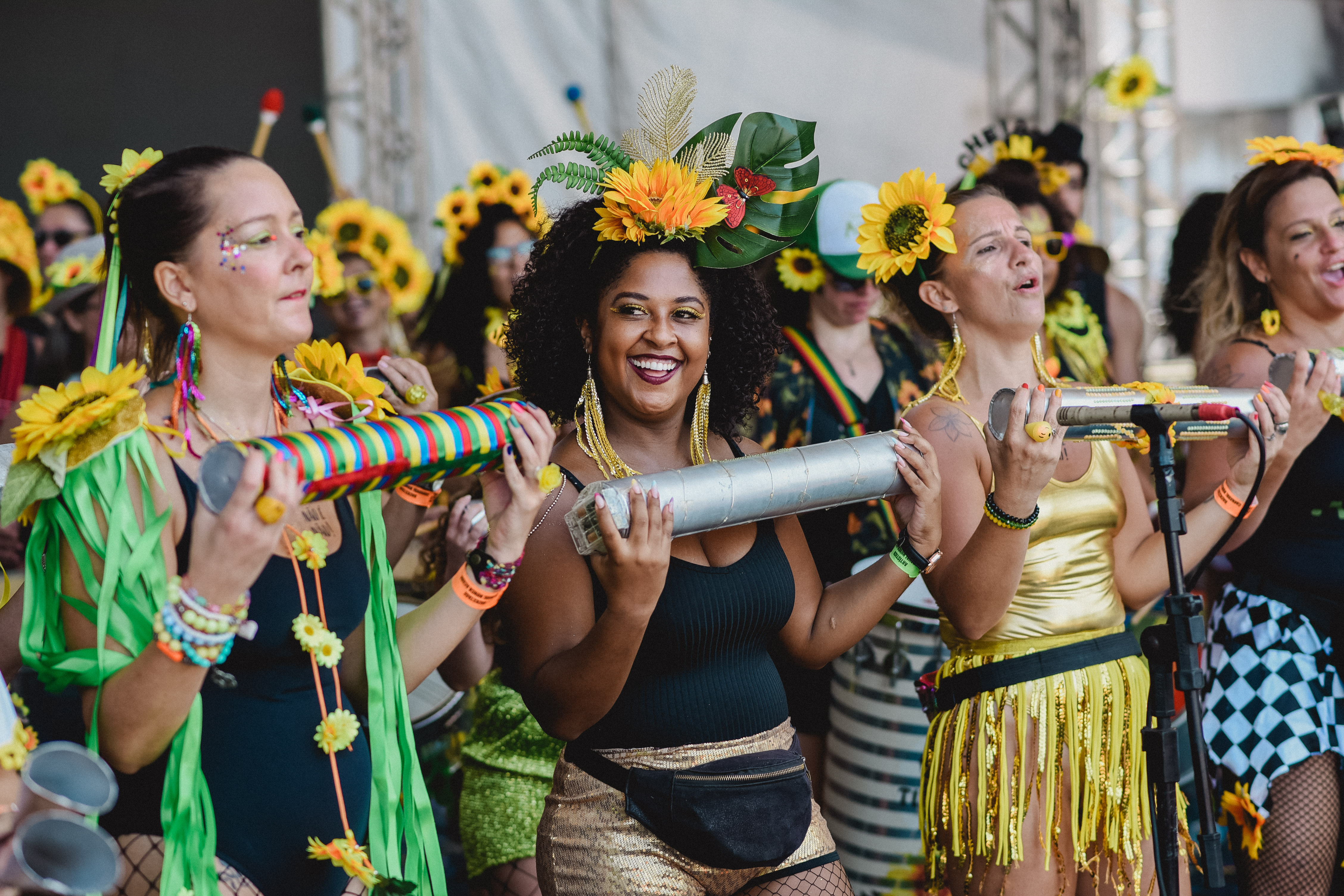 Mulheres instrumentistas do bloco Que Pena Amor com girassois na cabe&ccedil;a e tocando chocalho.
