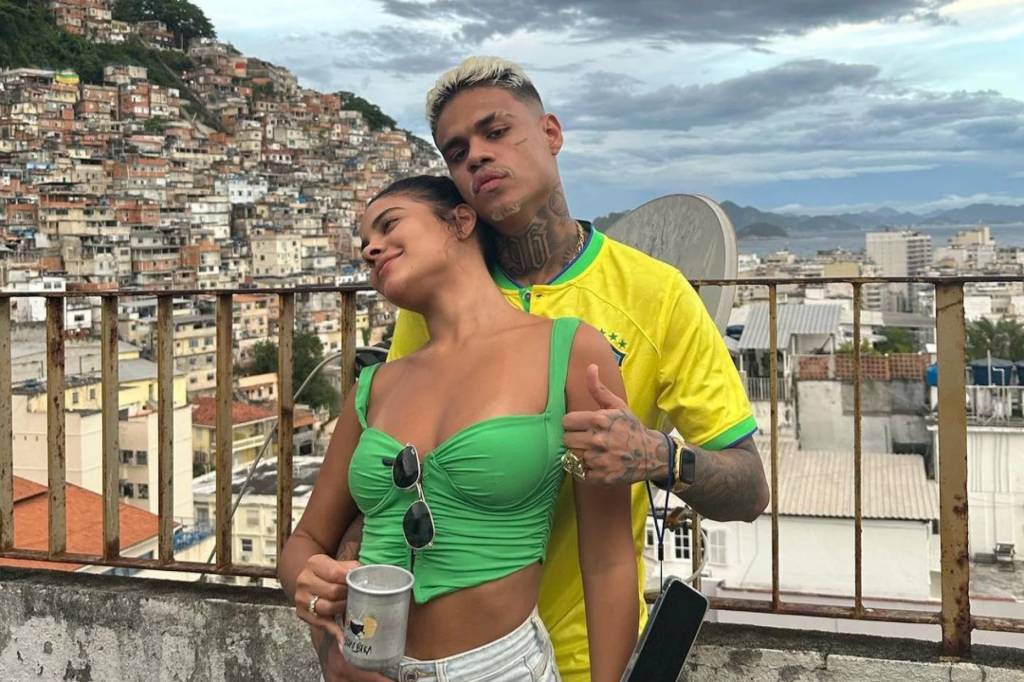 bella campos mc cabelinho MC Cabelinho usa camisa da seleção brasileira. Ele abraça Bella Campos por trás, e ela usa top verde e segura uma caneca na mão. Ao fundo, a vista para uma favela.