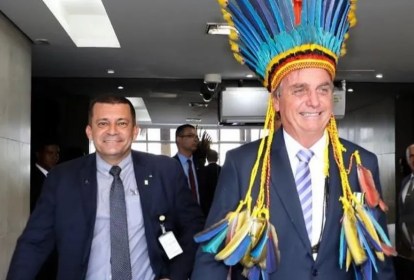 Foto mostra Bolsonaro usando um cocar indígena na cabeça e eu segurança ao lado, ambos usando paletó, camisa social e gravata