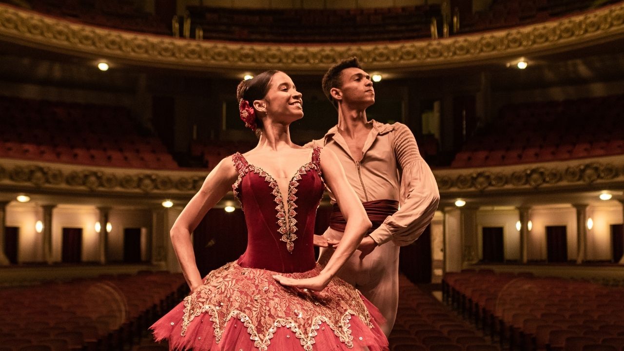 Jovem branca, de cabelo castanho preso em coque, vestida com roupa de balé vinho, posa fazendo pas-de-deux com jovem negro, com roupa em tons de marrom claro. Ao fundo, a parte interna do Theatro Municipal.