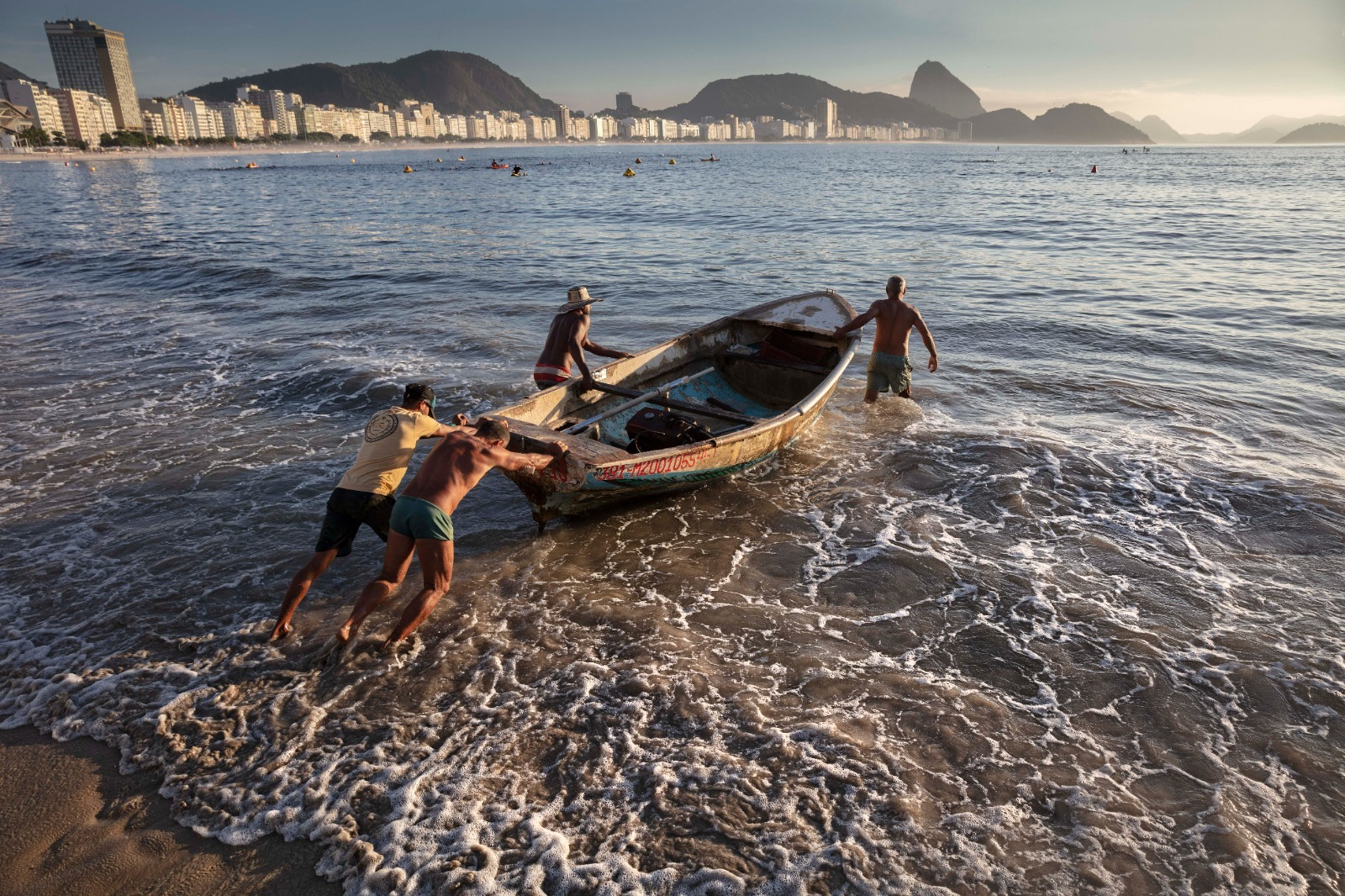 Quatro pescadores no posto 6 da Praia de Copacabana empurram um barco na areia da praia para o mar, tendo ao fundo o Morro da Urca e o P&atilde;o de A&ccedil;&uacute;car.