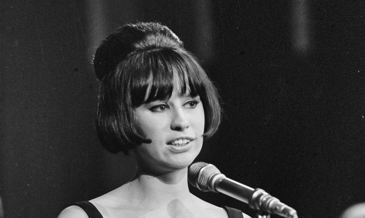 Foto em preto e branco de Astrud Gilberto, mulher branca de cabelo liso em corte chanel com grande coque na cabeça. Ela usa roupa com alças nos ombros e está diante de um microfone