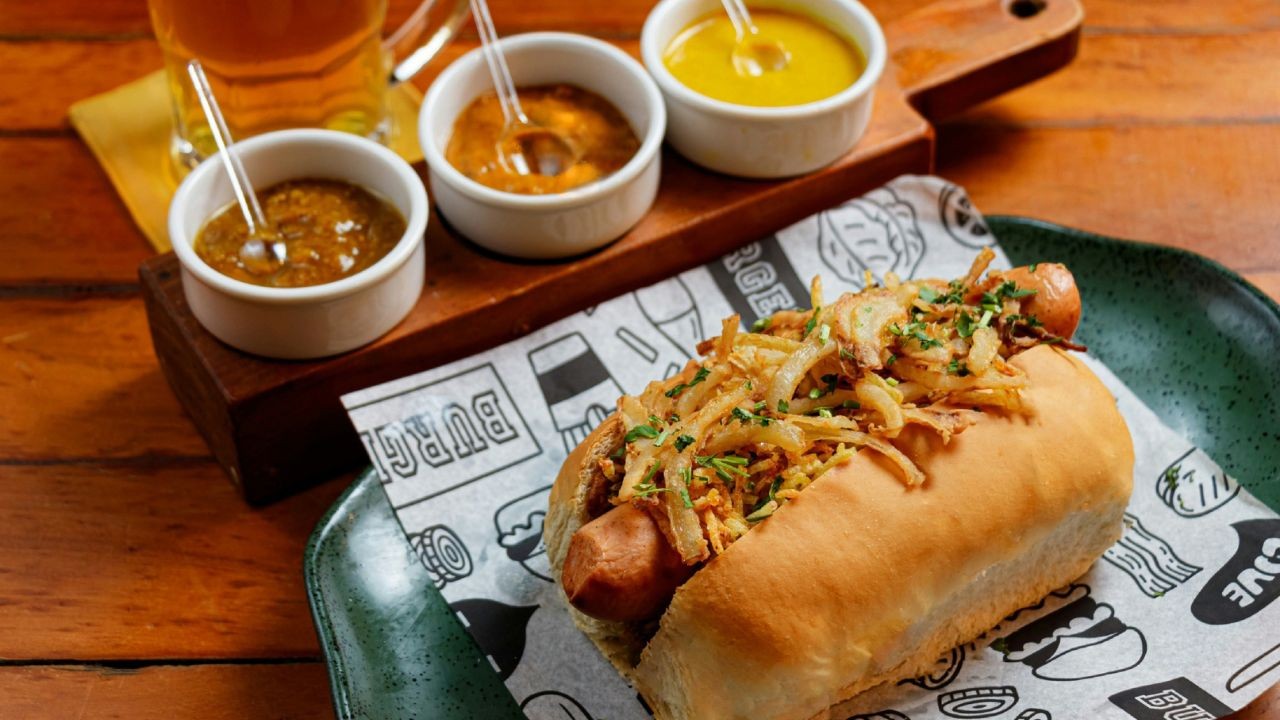 Um cachorro-quente gourmet com cebola caramelizada e salsinha picada, servido em papel estampado sobre um prato verde escuro. Ao fundo, três potes brancos com molhos e um copo de cerveja, tudo sobre uma mesa de madeira